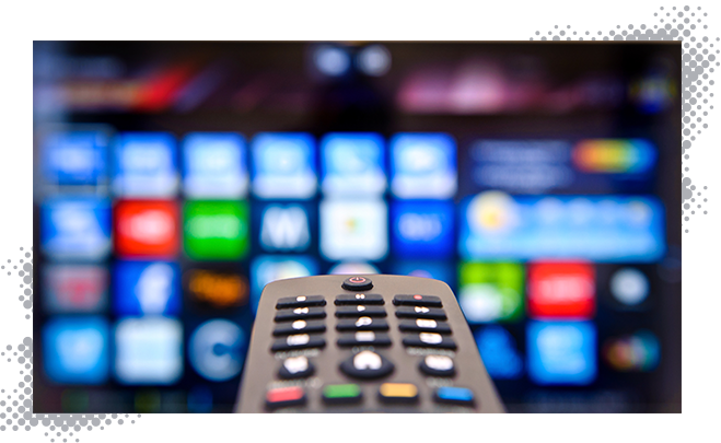 Close up of TV remote with multiple channel options on a TV in the background.