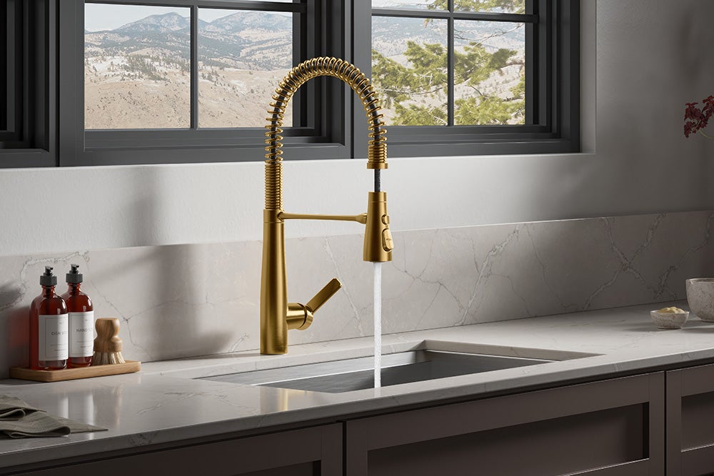 Gold Kohler Kitchen faucet sinks white marbeling counter top surrounding. Black window panes behind with mountain background through the window