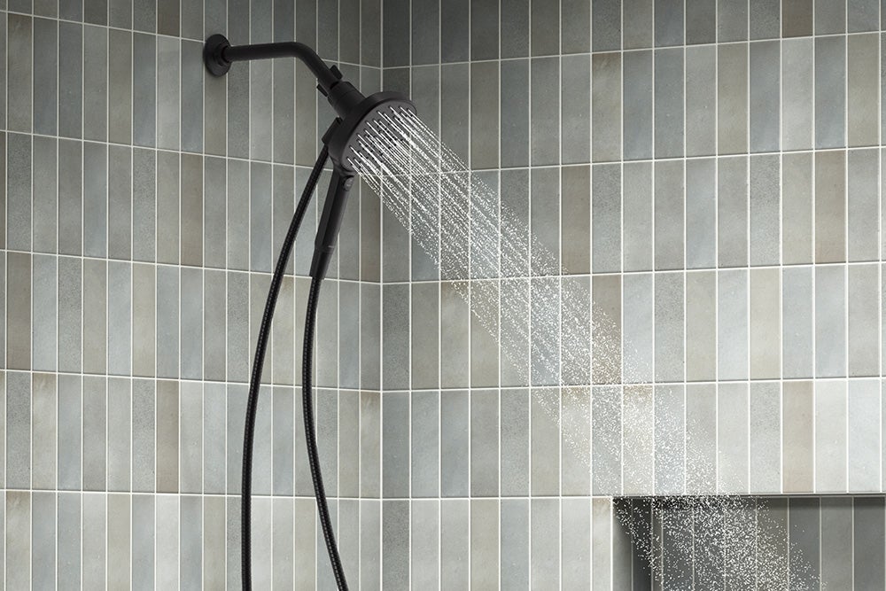 Black showerhead with running water in a mixed faint blue, grey, and brown bathroom tile.